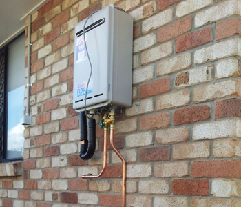 The image shows a gas hot water system mounted on a brick wall. The unit is grey with pipes connected to it, one black and one copper, indicating gas and water lines.