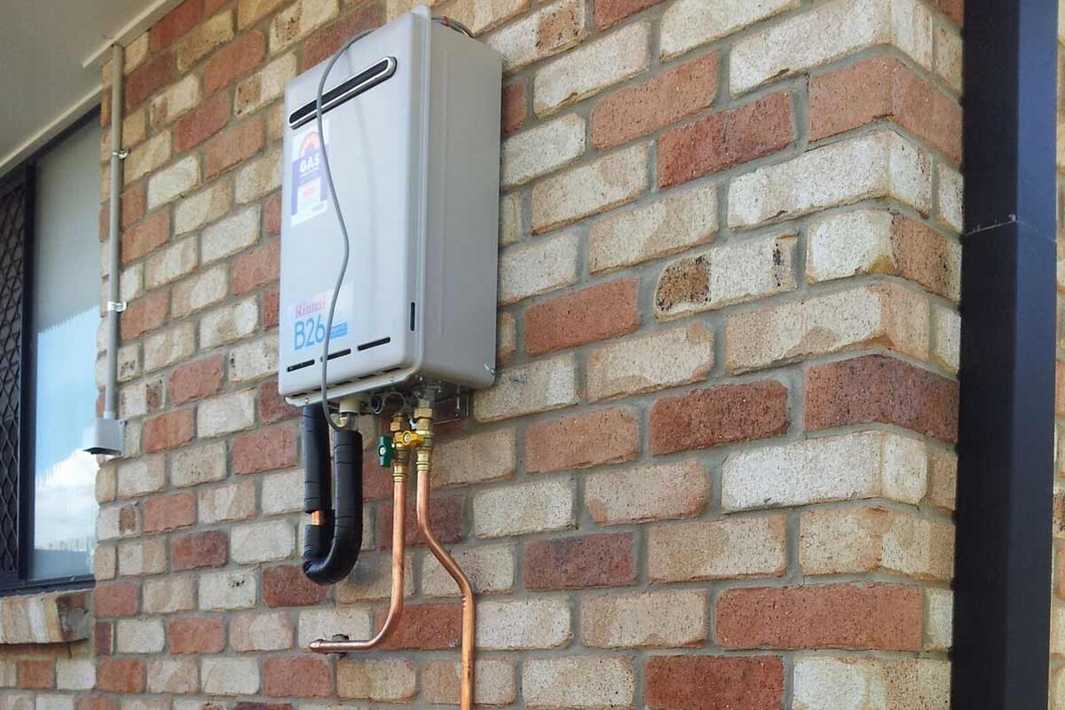 The image features a mounted gas hot water system installed on a brick exterior wall. The unit is compact and grey, with copper piping connected to it. The bricks are a mix of red and lighter hues.