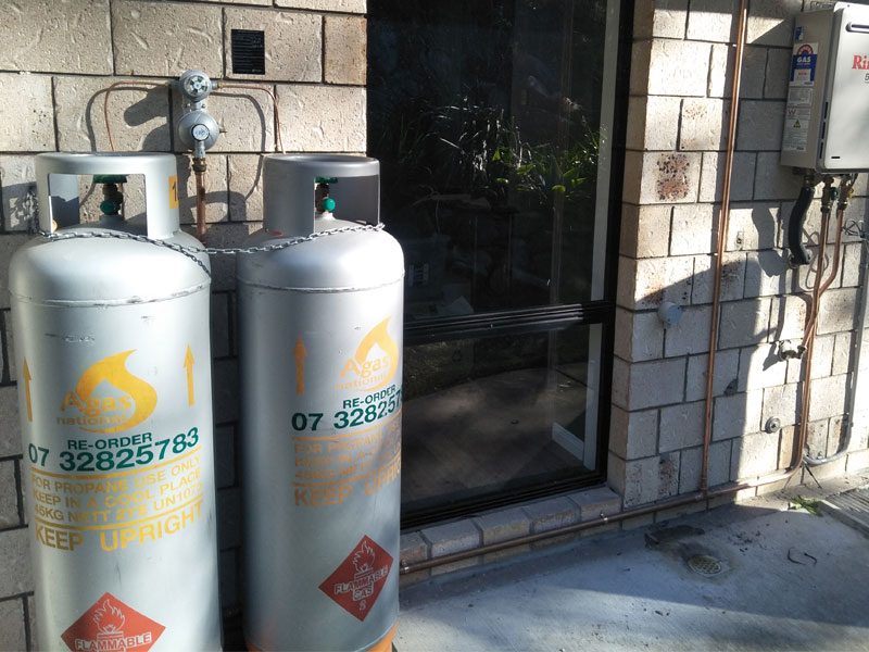 The image captures two large gas cylinders positioned beside a glass door. The cylinders are silver with warning labels and a visible re-order contact number. A gas meter is mounted on the wall, and some greenery is visible through the glass door, indicating an outdoor area.