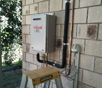 The image depicts a tankless gas water heater mounted on a wall. It features a metallic casing with the brand name visible on the front. Pipes connected to the heater indicate its installation for gas supply and water discharge. A ladder is positioned underneath the unit, suggesting ongoing installation or maintenance work.