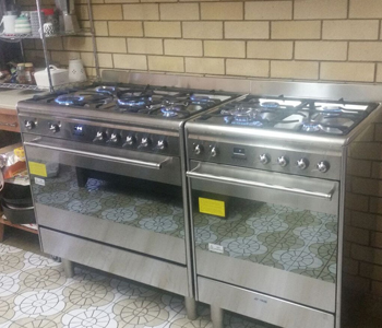 The image features two stainless steel gas cookers positioned side by side in a kitchen setting. Each cooker has multiple burners, with blue flames visible, indicating they are in use. The cookers have a modern design, with control knobs on the front and a sleek finish. The backsplash is made of light-coloured bricks, and the floor has distinctive patterned tiles.