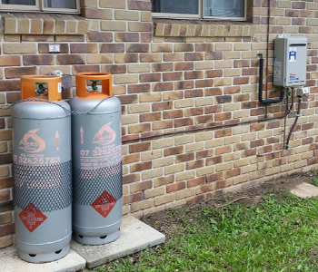 The image features two standard gas cylinders secured on concrete slabs beside a brick wall. A gas meter is mounted on the wall, with pipes extending from it.