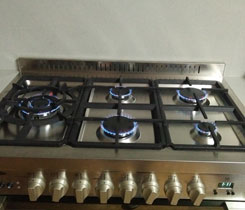 The image shows a stainless steel gas cooktop with five burners. The burners are ignited, displaying blue flames, and the control knobs are neatly arranged along the front of the cooktop.