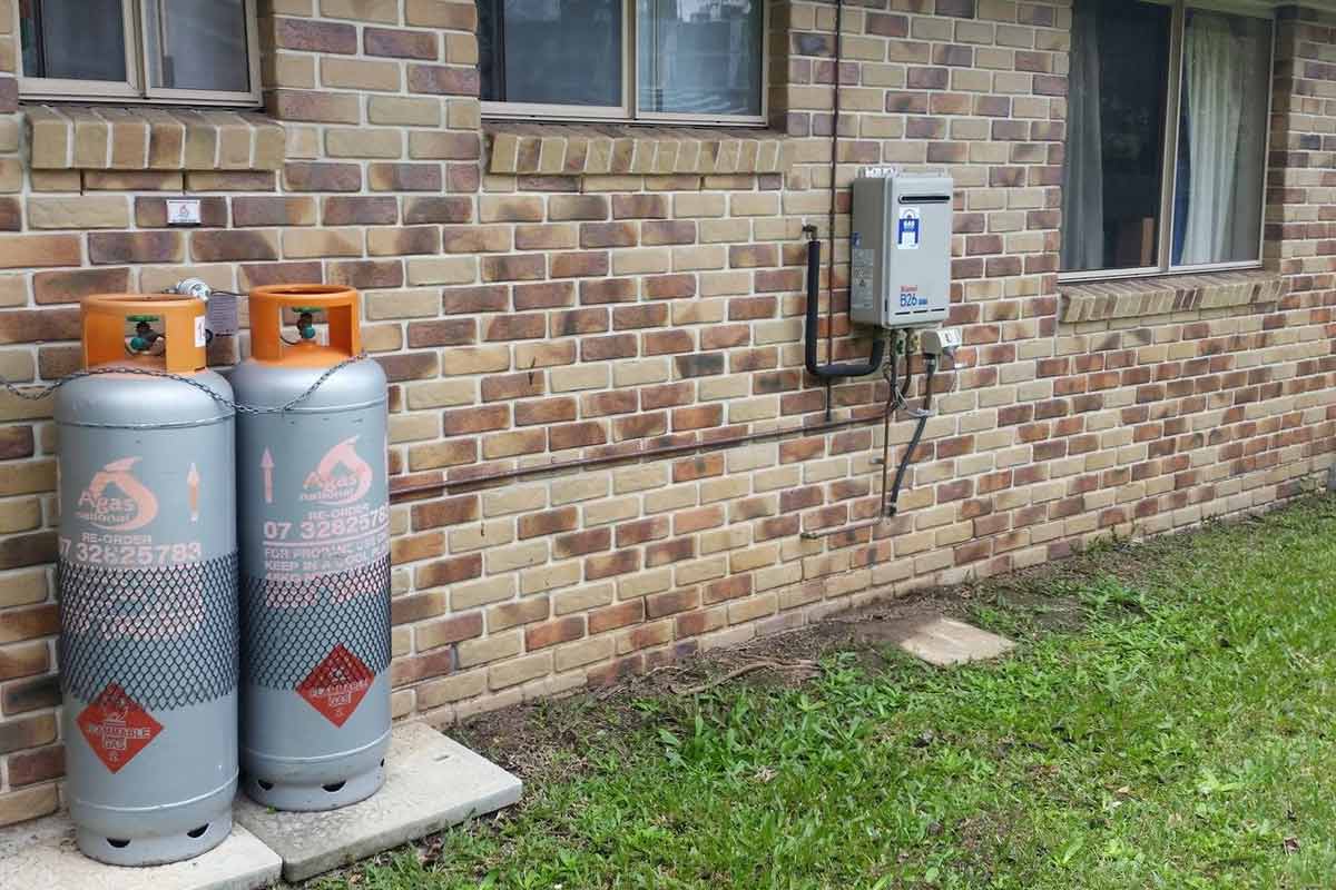 The image features two grey gas cylinders, prominently displayed at the side of a brick building. Each cylinder has a distinctive orange cap and safety markings. Adjacent to the cylinders, there is a gas meter box installed on the wall, which is connected to pipes running along the brick surface. The surroundings include a small patch of grass and a clear view of the brick texture of the building.