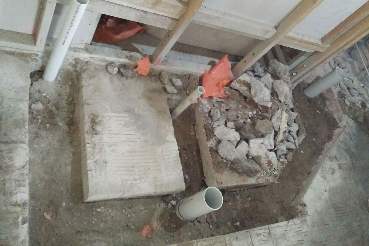 The image shows a construction site with exposed soil, rubble, and sections of white piping. There are piles of broken concrete and debris scattered around, indicating ongoing renovation or excavation work. Two vertical wooden beams are also visible, providing support in the area.