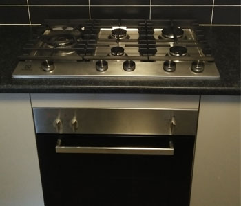 The image features a modern stainless steel gas cooktop with five burners and a sleek black oven below. The cooktop has various dials for adjusting heat and features a clean, contemporary design, set against a dark tiled backsplash.