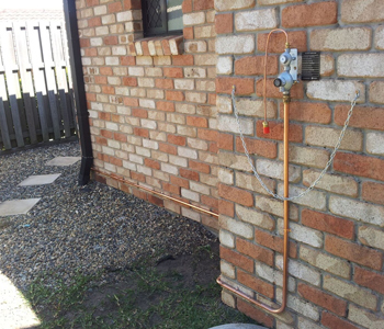 The image shows a gas installation on the exterior of a brick wall. Copper pipes are prominently displayed, leading to a gas meter with a shut-off valve. The area around the installation features a gravel path and grass.